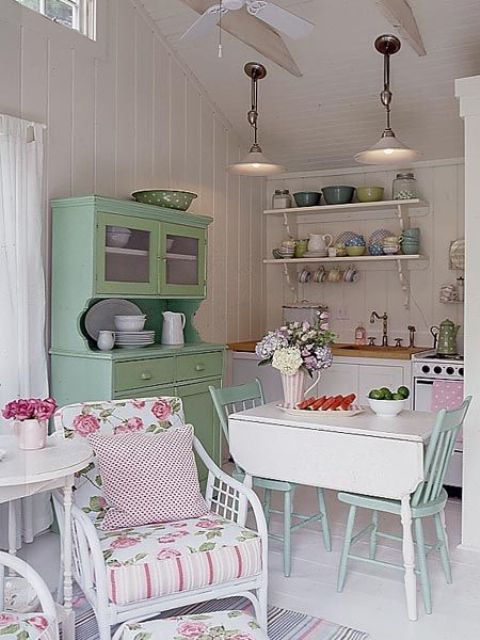 A neutral vintage kitchen with built in cabinets, a green buffet, a folding table and blue chairs, colored tableware and chairs with floral upholstery