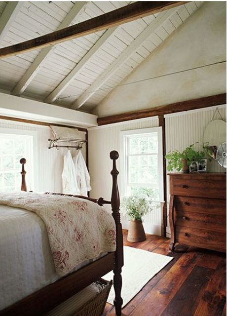 a cozy farmhouse bedroom with a white wooden ceiling plus beams, stained wooden furniture and some printed textiles 