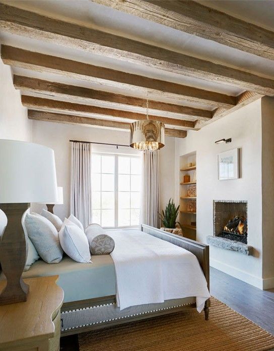 a neutral and chic farmhouse bedroom with wooden beams on the ceiling, a built-in fireplace and elegant furniture