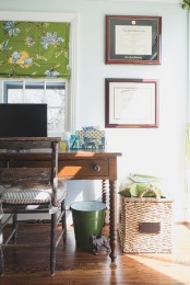 a cozy farmhouse home office with vintage wooden furniture, a bucket for trash and a basket for storage, a green floral curtain and some artworks on the wall