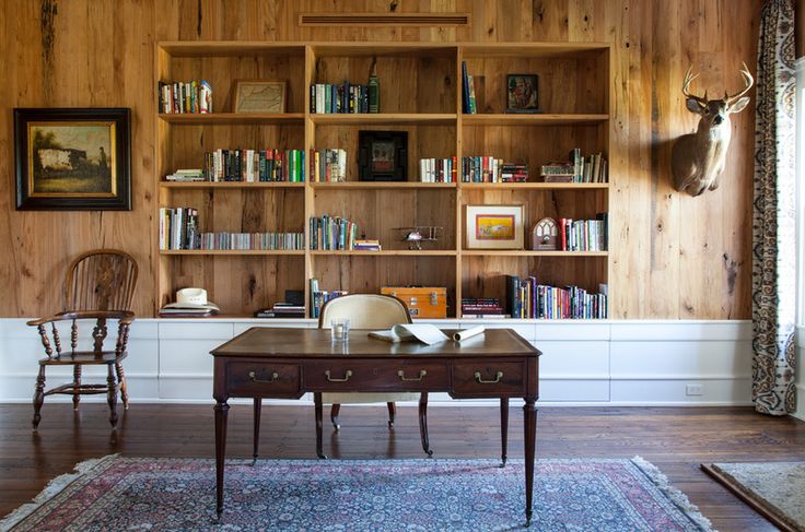 A farmhouse home office with a wood clad wall, built in bookshelves in a niche, a vintage desk and some chairs plus a bold rug