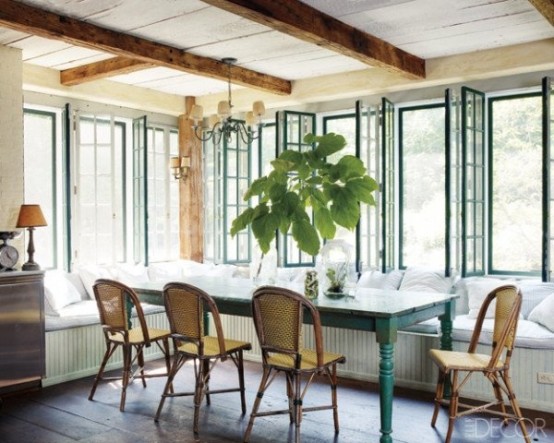 a farmhouse sunroom with a dining zone  with a long L-shaped window seat, a blue table and rattan chair plus wooden beams