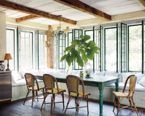 A farmhouse sunroom with a dining zone  with a long L shaped window seat, a blue table and rattan chair plus wooden beams