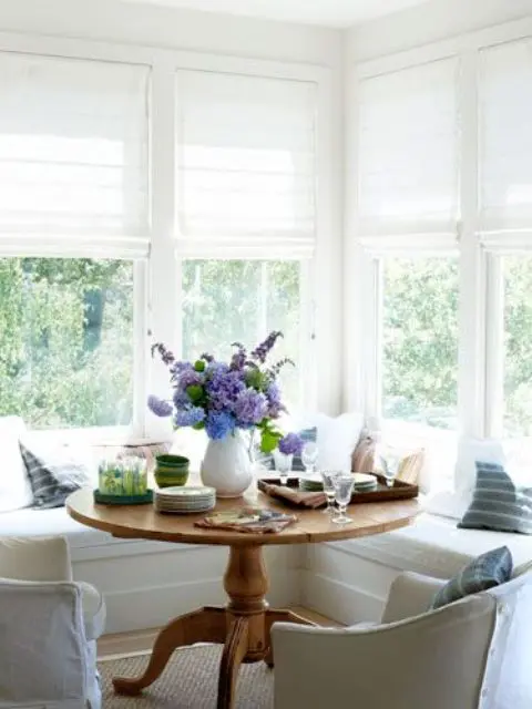 A neutral farmhouse sunroom corner with an L shaped window seat and a rustic vintage table for having breakfasts here