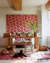 a creative boho chic home office with a wooden desk and stool, a comfy sofa for sitting, a pink wall hanging and a rug plus tropical leaves
