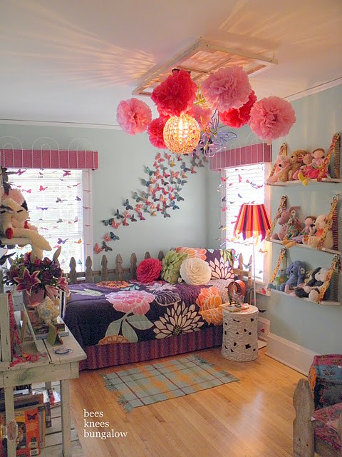 a colorful and whimsical kid's room with light blue walls, a purple bed with colorful bedding, colorful butterflies on the wall, paper pompoms and pendant lamps and colorful toys