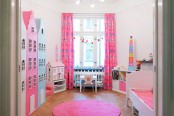 a colorful kid room with large dollhouses, a bed with pink bedding, a hot pink rug, a working space by the window with pink curtains