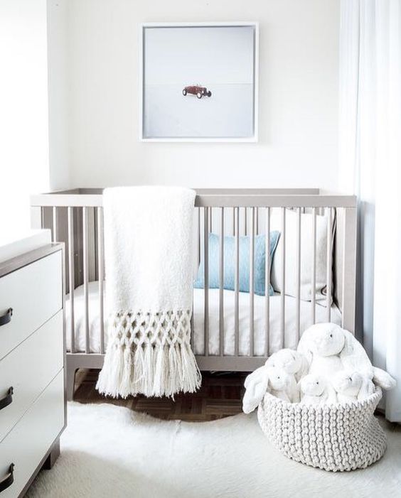 A white gender neutral nursery with white and grey furniture, white and blue bedding, toys and an artwork is serene and airy