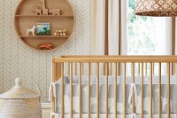 a gender neutral nursery with light-colored furniture, printed bedding, a basket and a round wooden shelf is a very chic space