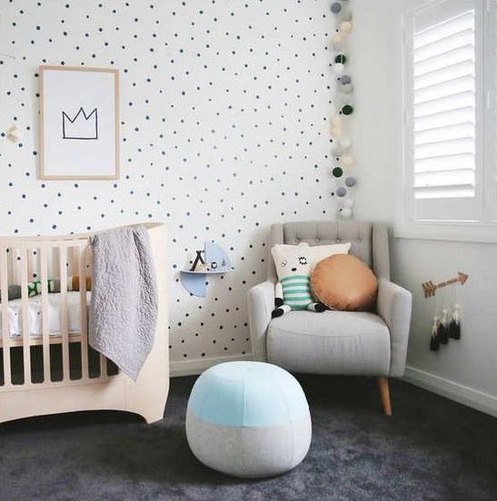 a gender neutral nursery with a spotted wall, a wooden crib, a grey chair and a blue ottoman and some plush and soft touches