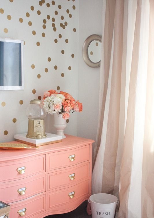 a cute and glam gold polka dot accent wall and a coral dresser with gold knobs, a gold tray and vase is a lovely and playful decor idea