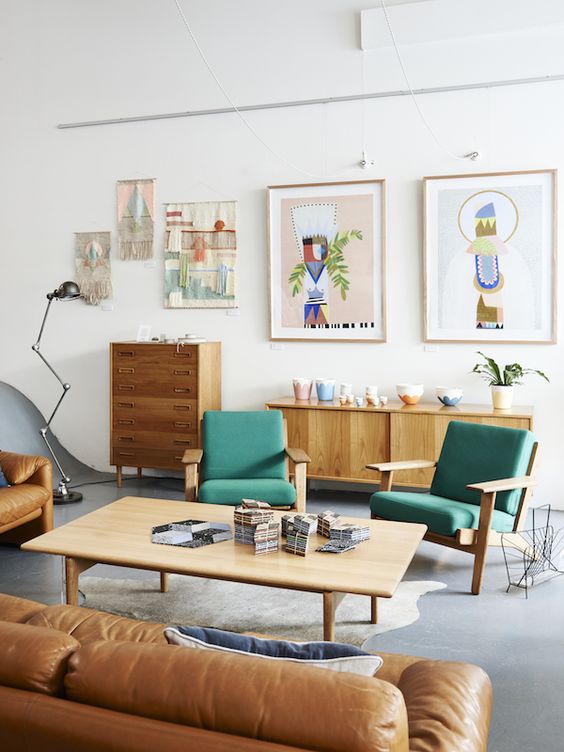 A bright mid century modern living room with stained furniture, green chairs, an amber leather sofa, a low coffee table, a gallery wall and beautiful potted plants