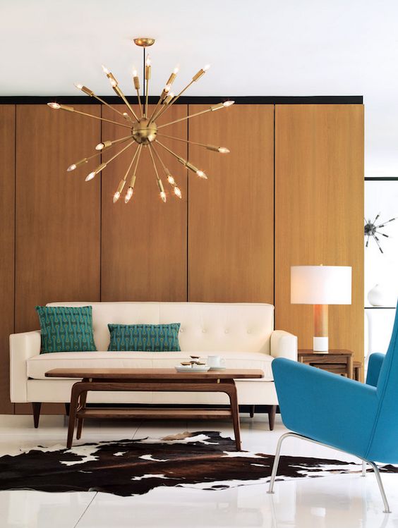 A mid century modern living room with a plywood wall, a creamy sofa, a turquoise chair, a low coffee table, a sunburst chandelier and a cowhide rug