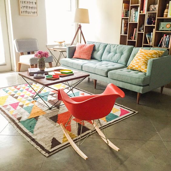 A bright mid century modern living room with an aqua sofa, bright pillows, a colorful printed rug, a coffee table and rocker chairs, a large bookcase and floor lamps