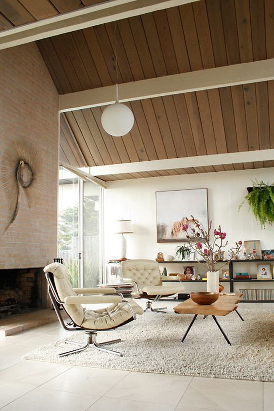 A neutral mid century modern living room with a large fireplace, creamy chairs, a glass coffee table, an open shelving unit and greenery is amazing
