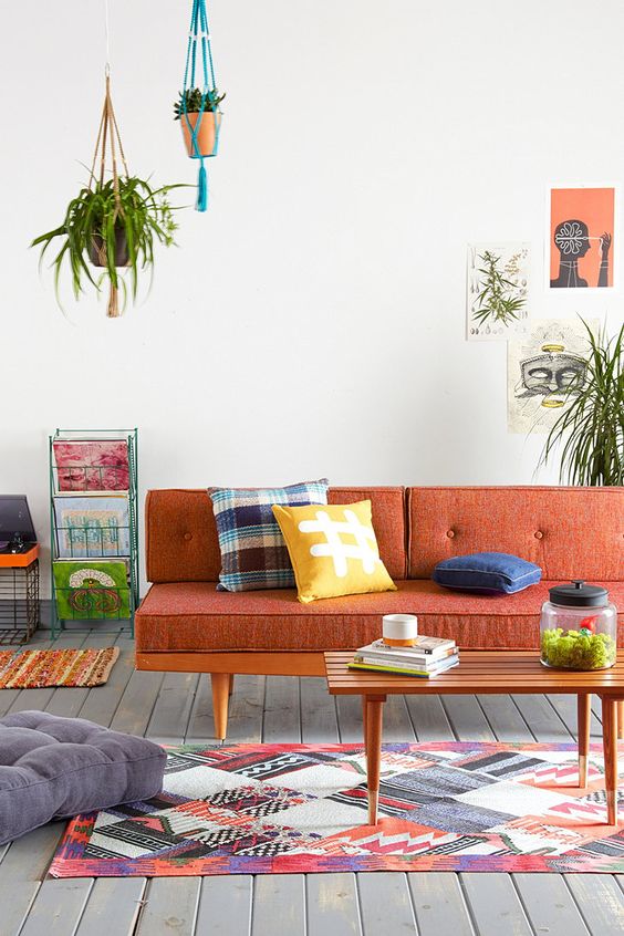 A mid century modern to boho living room with a rust colored sofa and bold pillows, a bright rug, potted greenery and shelving units with magazines