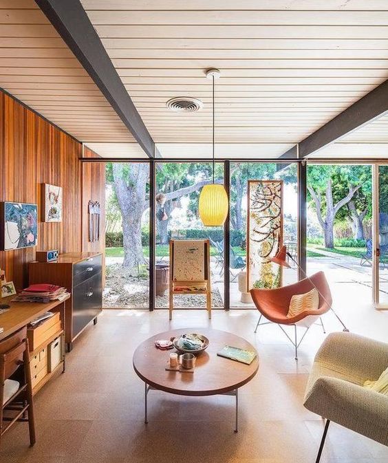A mid century modern living room with a wood clad wall and glazed ones, stained storage furniture and seating furniture, a low coffee table, cool artwork
