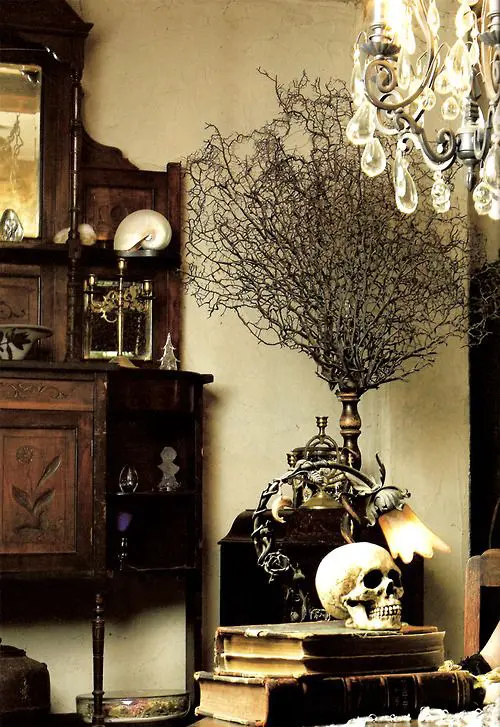 a refined Gothic home office with dark and heavy carved wooden furniture, a crystal chandelier, stacked books and a skull plus bold accessories