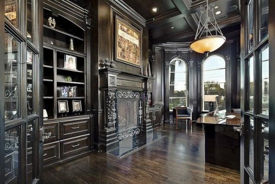 a dark vintage home office with heavy and dark furniture, a non-working fireplace, shelves and a desk, some chairs by arched windows