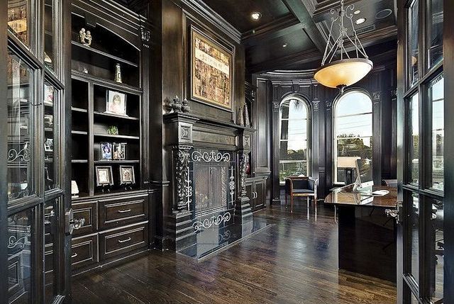 A dark vintage home office with heavy and dark furniture, a non working fireplace, shelves and a desk, some chairs by arched windows