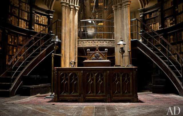 A Gothic library with bookshelves raising up to the ceiling, curved staircases, a heavy carved desk in the center and a chair   the whole space looks like out of a movie