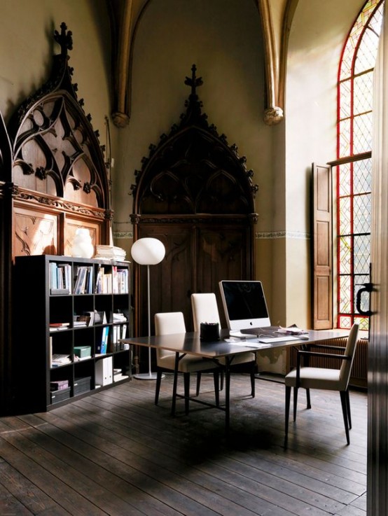 a sophisticated gothic home office with vintage carved wooden doors, a black shelving unit, a large desk, creamy chairs and a giant church-like window