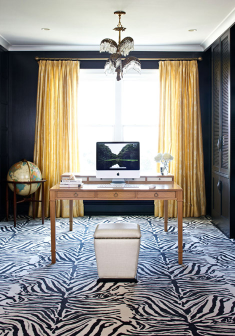 A chic home office with black walls and a built in storage unit, a light stained desk and a white leather stool, a zebra print rug plus a large vintage globe