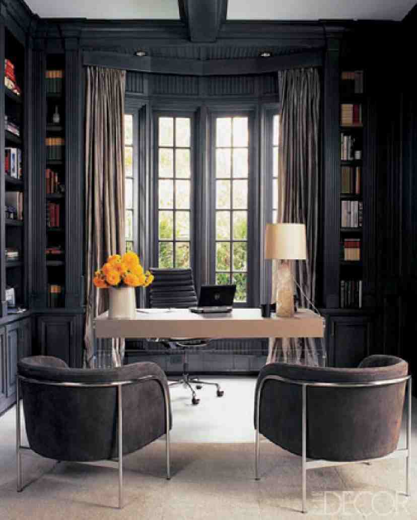 A sophisticated moody home office with black walls and built in bookcases, a light stained and sleek desk, brown chairs and a cool bay window