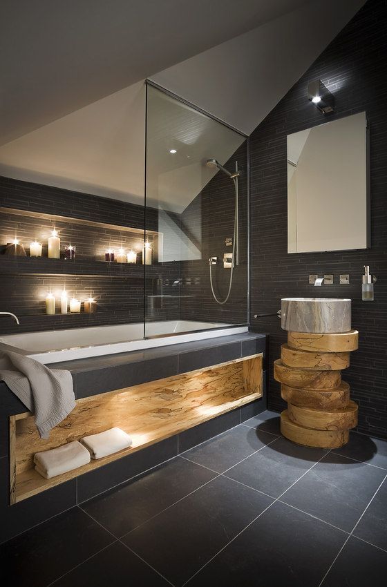 A dark home space with a tub and niches with candles over it, a unique vanity of tree slices, a wooden niche for towels and built in lights welcomes in