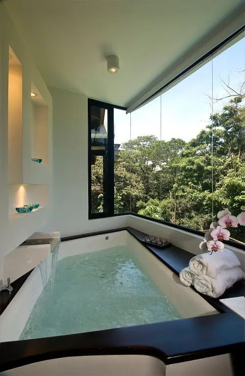 a contemporary bathroom with a glazed wall, a large hot tub with a waterfall and built-in niches and shelves is a lovely space to relax while enjoying the views