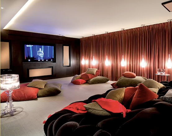 A contrasting living room with a built in screen, red curtains, catchy pendant lamps, a black sofa and piles of colorful pillows