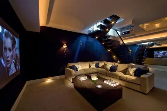 a pretty living room turned into a home theater, with a large screen, a corner sofa, a brown pouf and built-in lights around