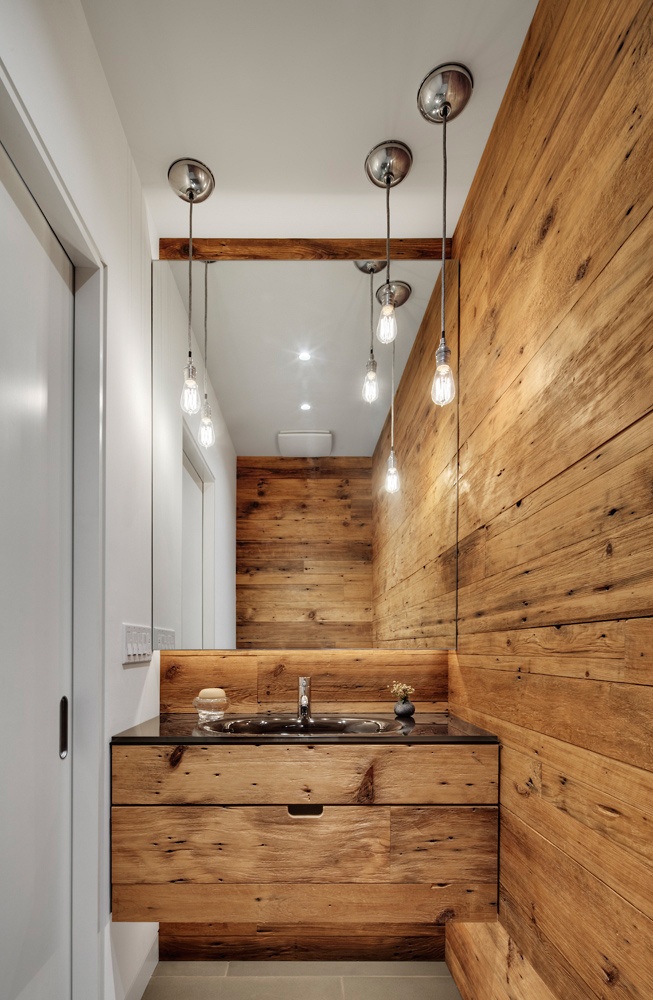 A modern bathroom with a chalet feel   a light stained wooden wall, a matching built in vanity, a large mirror and bulbs hanging down