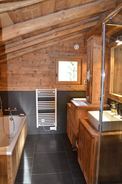 A small chalet bathroom with large scale tiles and light stained wood, with a small window, wooden furniture and a tub clad with wood