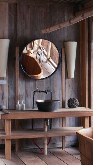 A vintage inspired chalet bathroom clad with dark wood, with light stained wooden pieces and a round mirror, a vintage metal sink and a faucet