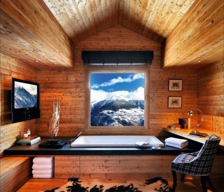 A stylish and cool chalet bathroom clad with light stained wood, with a view, a TV, a chair, towels, lamps and a blanket