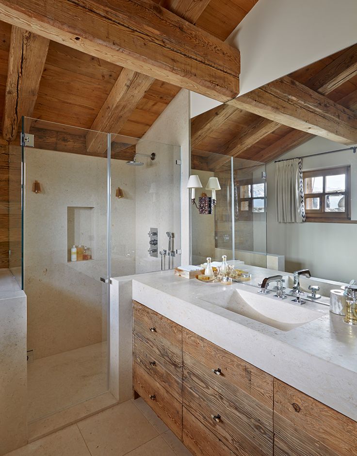 A light filled chalet bathroom with windows, with a light stained wood ceiling with beams and a matching vanity, neutral stone on the floor, in the shower