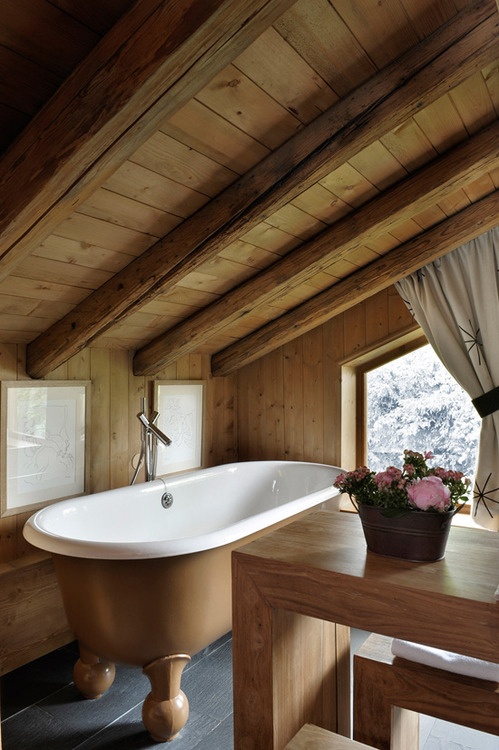 A light stained wood attic chalet bathroom with a window, a clawfoot bathtub, a light stained table and neutral textiles