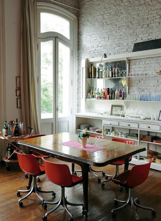 an eclectic dining space with a whitewashed bricl wall, a large buffet and shelves, a refined wooden table and modern red chairs