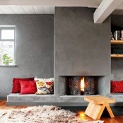 a contemporary space with a concrete clad fireplace and seating spaces with pillows plus a wooden stool for enjoying warmth here