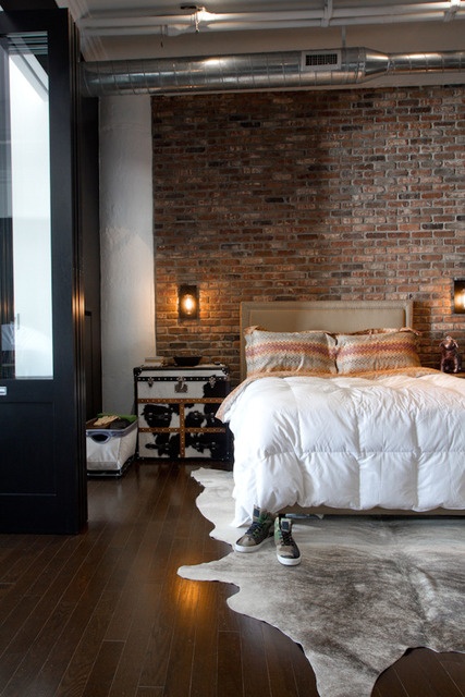 a stylish bedroom with a red brick accent wall, a bed with neutral bedding, exposed pipes under the ceiling, a chest with metal as a nightstand and a cozy rug
