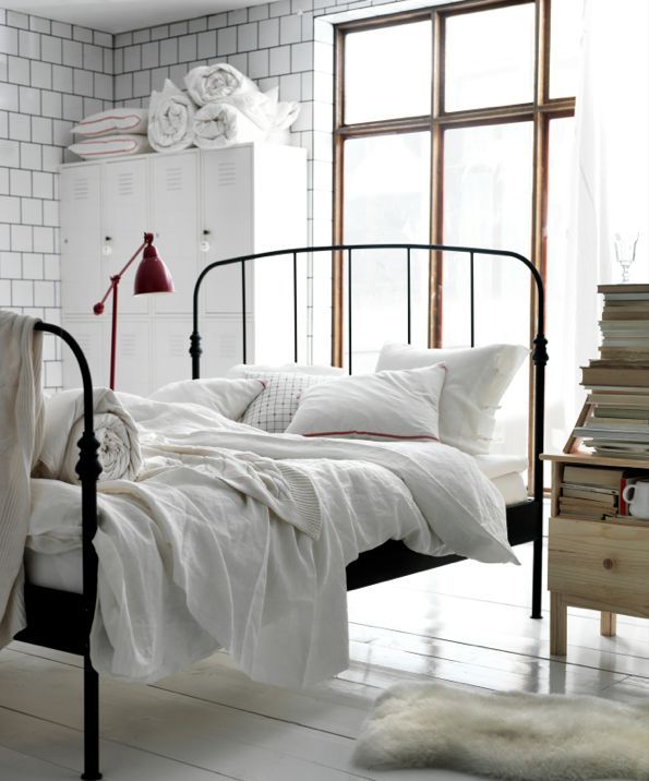 A light filled bedroom with tiled walls, a metal bed with neutral bedding, a white metal file cabinet for storage, a wooden nightstand with books