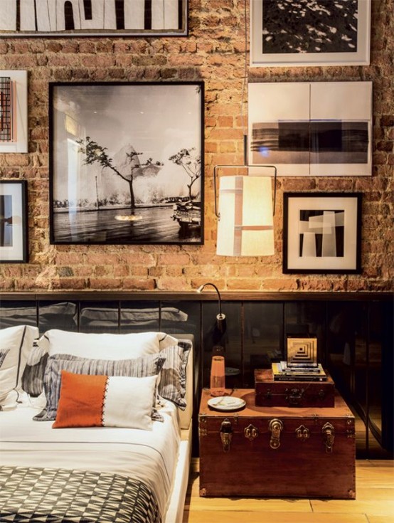 an industrial bedroom with brick walls, wooden paneling, a gallery wall, a metal bed with printed bedding, a chest for storage and some books is cozy and welcoming