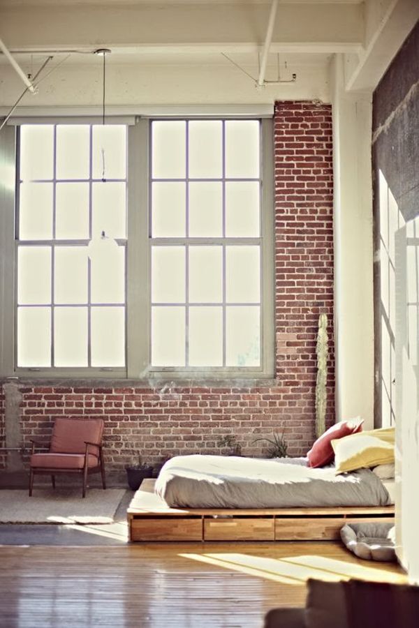 An industrial bedroom with brick walls, double height windows, a wooden bed with storage drawers and neutral bedding, pipes and metal on the ceiling