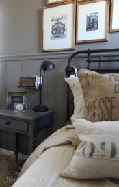 a vintage industrial bedroom with grey walls, a metal bed with printed bedding, a grey nightstand, a black table lamp and a gallery wall