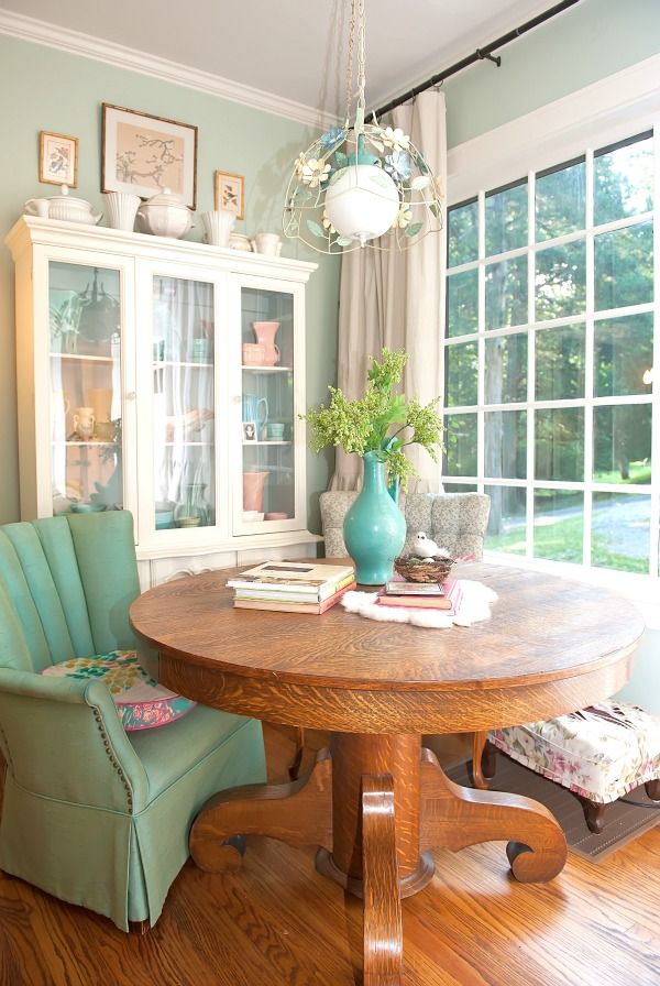 A chic vintage dining space with a glazed wall, a large vintage buffet, a stained round table, a mint green upholstered chair and a floral stool plus a chic chandelier