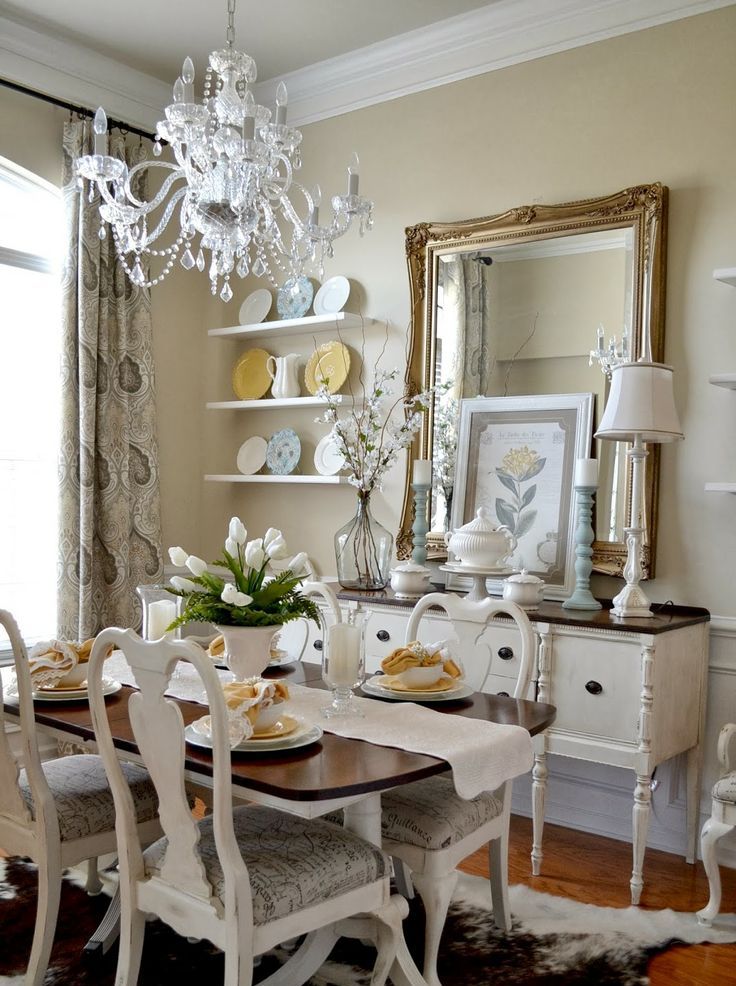 A neutral vintage dining room with tan walls, wall mounted shelves, a mirror and a buffet, a table with a stained tabletop, white vintage chairs and a white chandelier