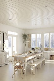 a vintage all-white dining room with extensive glazing, a vintage white table with chairs and benches, a buffet and a console table plus some greenery