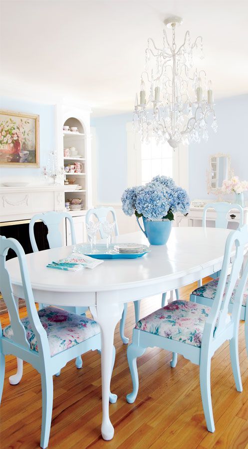 A chic vintage dining space with dusty blue walls, a non working fireplace, a white rounded table, blue chairs with floral upholstery and a beautiful white crystal chandelier
