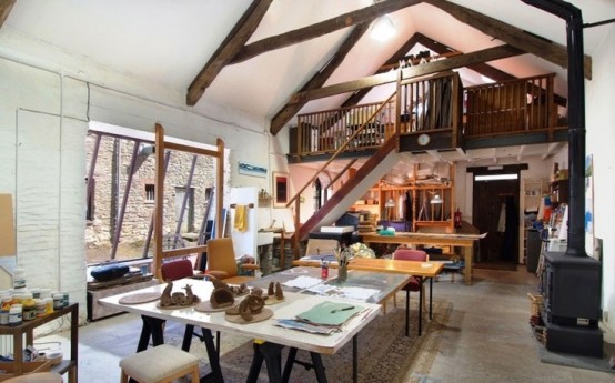 a large attic home studio with a hearth, several large tables for creating, a large window and dark-stained wooden beams that create a mood